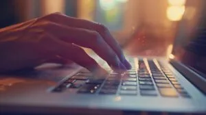 Pessoa limpando teclado de MacBook com pincel pequeno, iluminada por luz natural suave.